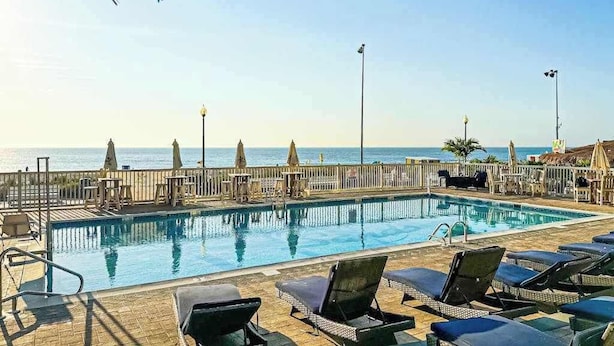 Uma piscina com espreguiçadeiras e guarda-sóis, um deck de madeira e vista para o oceano.