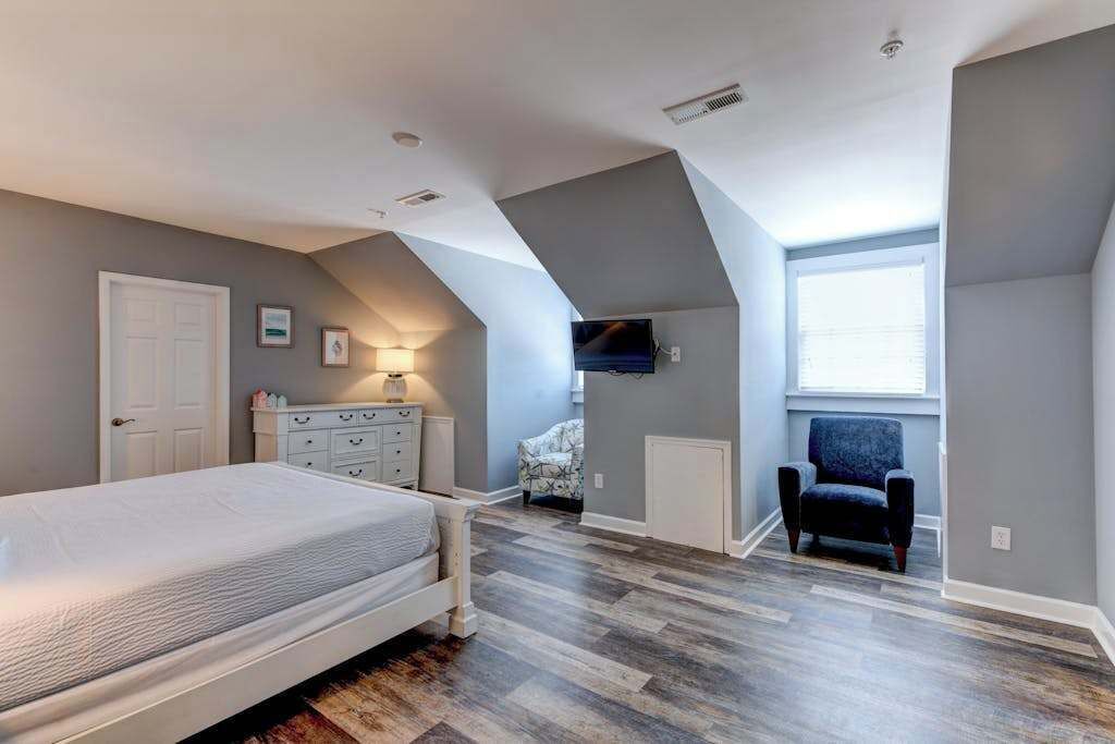 Ein Schlafzimmer mit Bett, Kommode, Stuhl und einem an der Wand befestigten Fernseher.