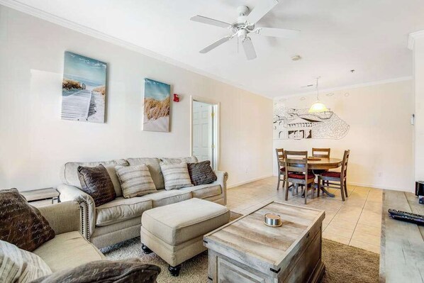 A living room with a sofa, ottoman, coffee table, dining table with chairs, ceiling fan, and artwork on the walls.