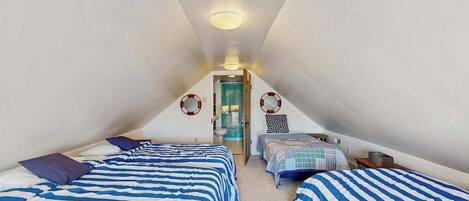 Ein quadratisches Schlafzimmer im Dachgeschoss mit zwei Betten, einer Tür zu einem Badezimmer und einer Deckenleuchte.