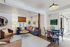 A modern living room with a sofa, coffee table, and dining area. A sign reads 'SLOW NO WAKE.' - Town of Lewes house w/deck, balcony, close to beach & restaurants (Lewes)