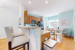 A modern kitchen with wooden cabinets, a granite countertop, and a dining area with a table and chairs. - Rehoboth Crossing townhome with pool, trails, EV charger, gym, balcony (Rehoboth Beach)