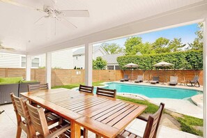 Un patio techado con mesa y sillas de madera, una piscina y mobiliario de exterior.