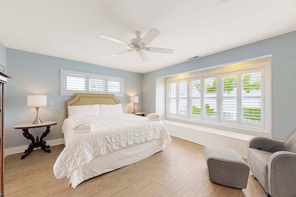 A bedroom with a bed, a ceiling fan, a window with blinds, and a nightstand with a lamp.