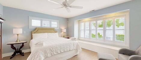A bedroom with a bed, a ceiling fan, a window with blinds, and a nightstand with a lamp.