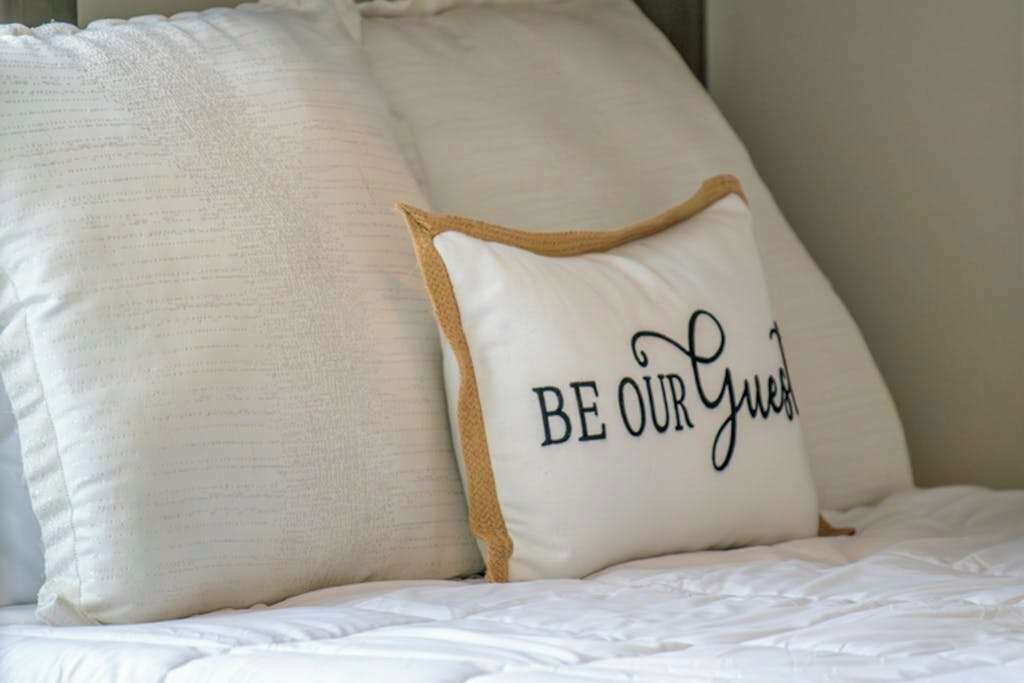 Una cama con ropa blanca y una almohada con el texto 'BE OUR Guest'.