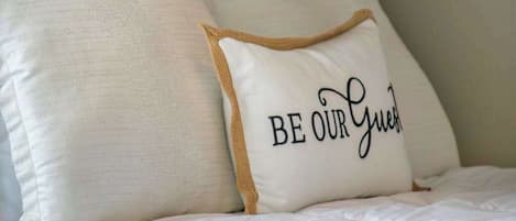 Una cama con ropa blanca y una almohada con el texto 'BE OUR Guest'.