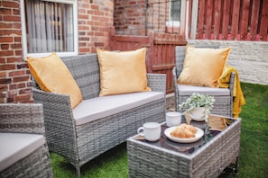 Wicker furniture with cushions, a table with a plant and a plate of food, and a brick wall in the background. - Modern Escape Oasis | Free Parking | 14PPL (Sheffield)