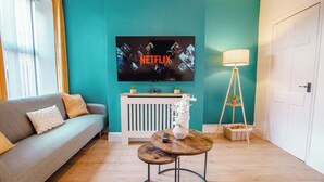 Ein Wohnzimmer mit einer Couch, einem an der Wand montierten Fernseher, einer Lampe und einem kleinen Tisch mit einer Vase und einer Fernbedienung.