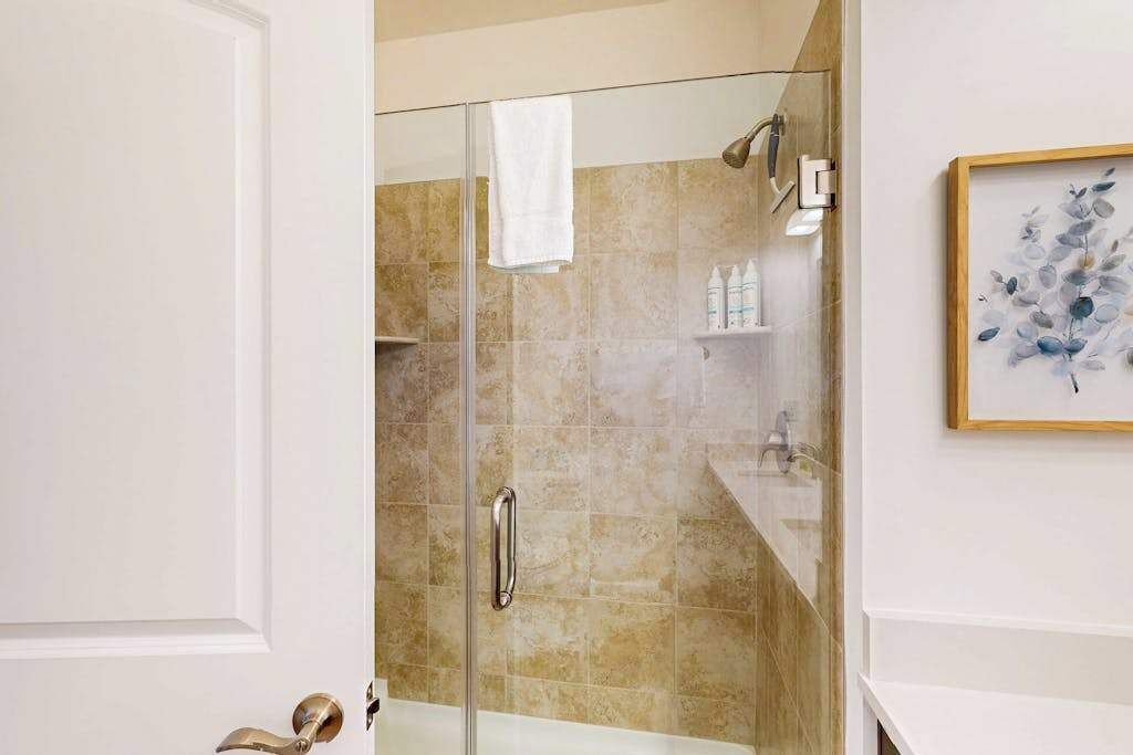 A glass-enclosed shower with a white towel hanging on the door handle, beige tiled walls, and a framed artwork on the wall.