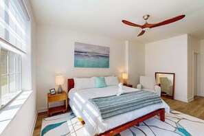 Un dormitorio con cama, dos mesitas de noche, ventilador de techo, un cuadro en la pared y ventana.