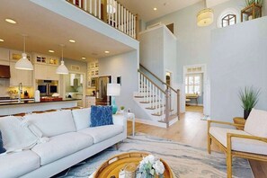 A spacious living area with a white sofa, a wicker chair, a staircase, and a kitchen in the background. - Arbor-Lyn house w/shared pool, deck, patio, ping-pong table, grill (Rehoboth Beach)
