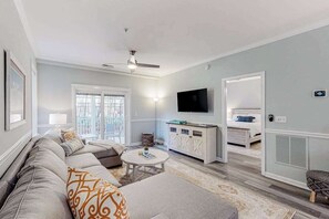 A living room with a sofa, a coffee table, a TV, and a bed in the adjacent room. - The Tides condo with pool access, covered patio, washer/dryer & central AC (Rehoboth Beach)