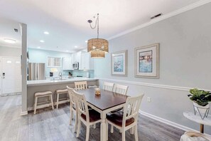 A dining area with a wooden table and chairs, a hanging light fixture, and framed artwork on the walls. - The Tides condo with pool access, covered patio, washer/dryer & central AC (Rehoboth Beach)