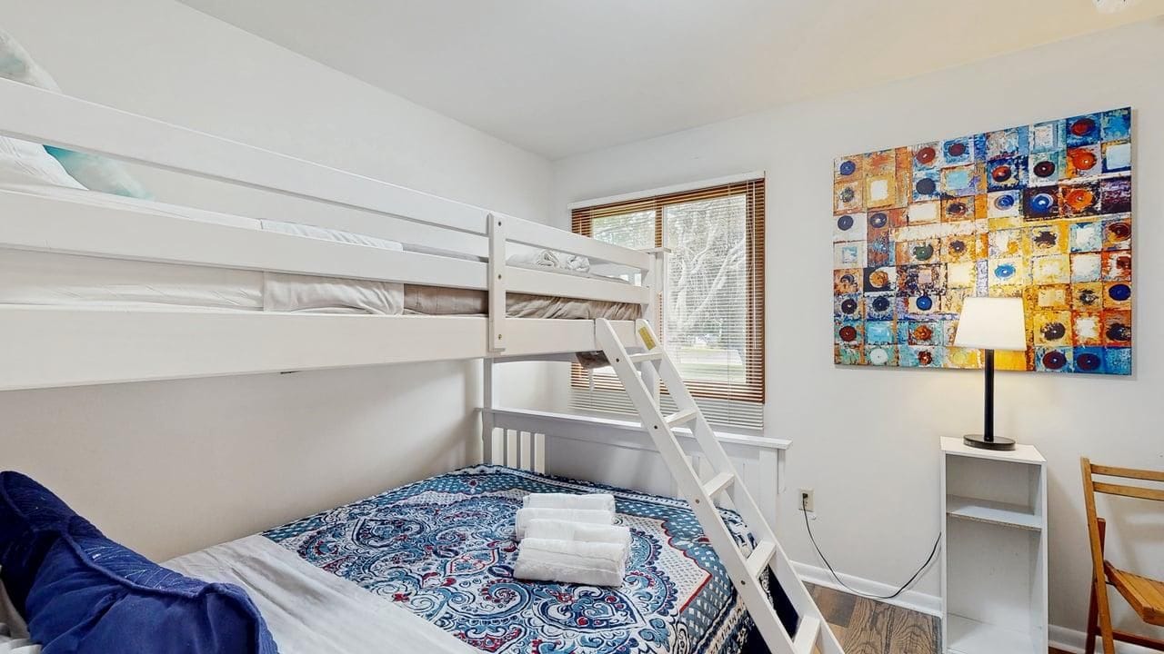 A bunk bed room with a ladder, a patterned bedspread, a wall art piece, and a bedside lamp.