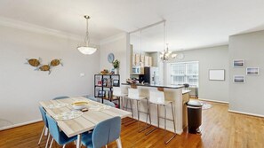 A modern kitchen with white cabinets, a dining area with a wooden table and blue chairs, and a visible trash bin. - Rehoboth Crossing townhouse with pool, gym, balcony, grill, W/D, bikes, & patio (Rehoboth Beach)