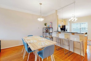 A dining area with a wooden table, blue chairs, a kitchen in the background, and pendant lighting. - Rehoboth Crossing townhouse with pool, gym, balcony, grill, W/D, bikes, & patio (Rehoboth Beach)