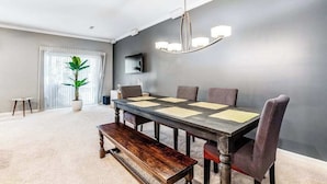 Un comedor con una mesa larga, sillas, un banco, una araña, una planta en maceta y un televisor.