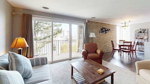 Una sala de estar con sofá, sillón, mesa de centro, mesa de comedor, sillas y un balcón con cortinas.