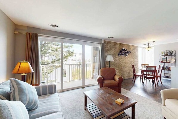 Ein Wohnzimmer mit Sofa, Sessel, Couchtisch, Esstisch, Stühlen und einem Balkon mit Vorhängen.