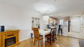 A dining area with a white table and chairs, a fireplace, and a kitchen in the background. - The Palms water-view condo near beach with outdoor pool, streaming, W/D, & AC (Rehoboth Beach)