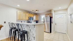 A kitchen with wooden cabinets, a stainless steel refrigerator, and a bar area with stools. - The Palms water-view condo near beach with outdoor pool, streaming, W/D, & AC (Rehoboth Beach)