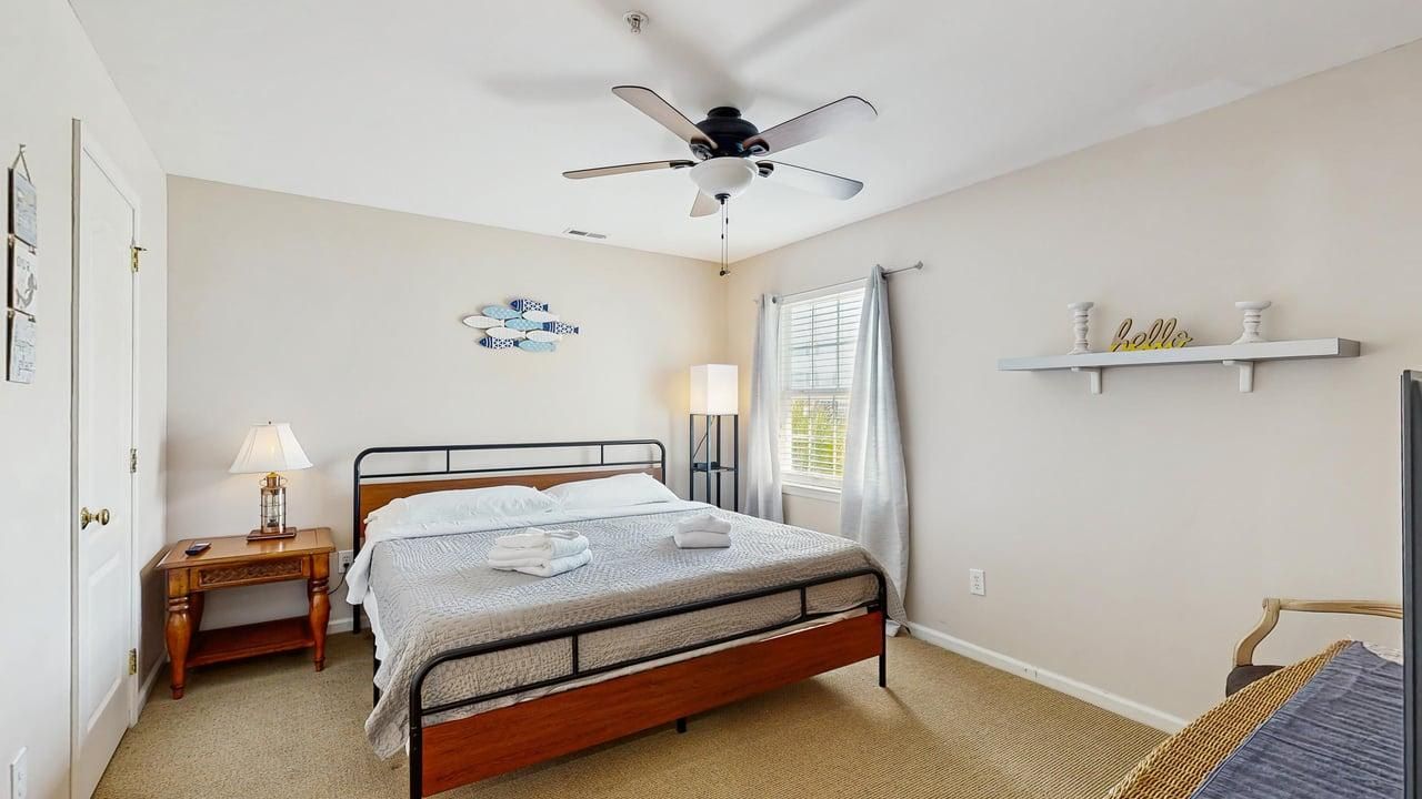 Une chambre à coucher comprenant un lit, une table de chevet, un ventilateur de plafond, une fenêtre avec des rideaux et une étagère ornée d’objets décoratifs.