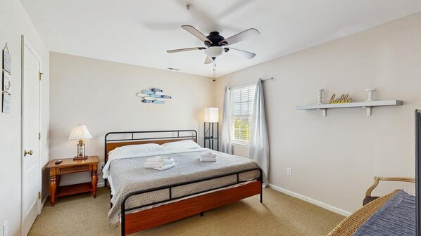 A bedroom with a bed, a nightstand, a ceiling fan, a window with curtains, and a shelf with decorative items. - The Palms water-view condo near beach with outdoor pool, streaming, W/D, & AC (Rehoboth Beach)