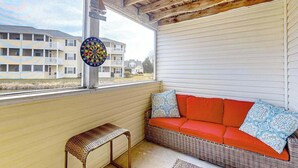 A room with a wicker sofa, a small bench, a dartboard, and a view of a building with balconies. - The Palms water-view condo near beach with outdoor pool, streaming, W/D, & AC (Rehoboth Beach)