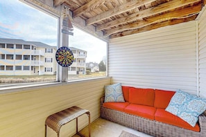 A room with a wicker sofa, a small bench, a dartboard, and a view of a building with balconies.