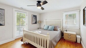 Un dormitorio con una cama blanca, ventilador de techo, radiador, mesita de noche y ventana con persianas.