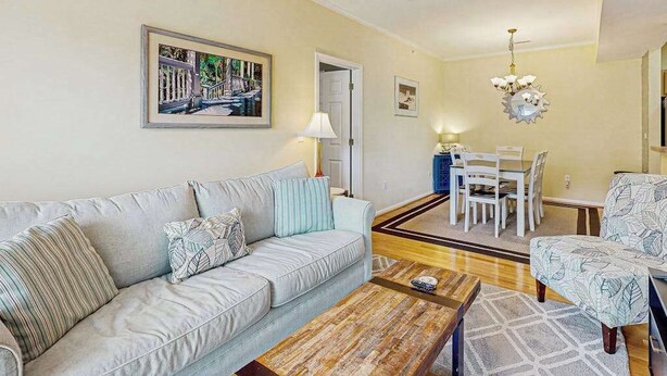 A living room with a sofa, coffee table, patterned armchair, dining table with chairs, and a chandelier. - Eagles Landing comfy condo with pools and tennis courts - 1 mile to beach (Rehoboth Beach)
