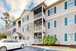 A multi-story building with balconies, a parking lot, and a small tree. - Spacious Creekwood condo with shared pool, Balcony, W/D & Free WIFI (Rehoboth Beach)