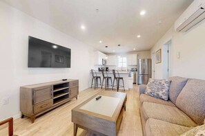 Una sala de estar moderna con sofá, mesa de centro, televisor y una cocina al fondo.