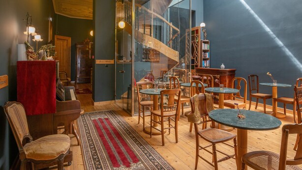 Ein gemütliches Zimmer mit einem Klavier, Holzsesseln, einer Glasstairnase und einem Bücherregal.