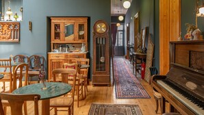 Ein Zimmer mit einem hölzernen Klavier, einer alten Standuhr, einem Essbereich mit Stühlen und einem Tisch sowie einem Flur mit einem gemusterten Teppich.