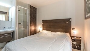 A bedroom with a bed, bedside lamps, a nightstand, a wooden headboard, a glass shower enclosure, and a bathroom with a sink and mirror. - Two bedroom Croisette Apartment 61 (Cannes)