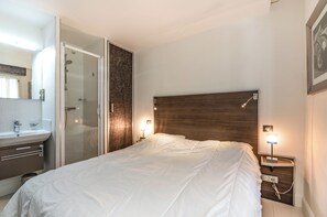 Ein Schlafzimmer mit Bett, Nachttischlampen, einem Nachttisch, einem hölzernen Kopfteil, einer Duschkabine aus Glas und einem Badezimmer mit Waschbecken und Spiegel.