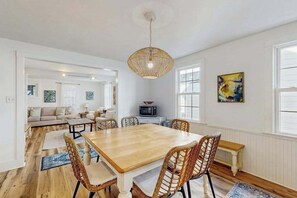 A dining area with a wooden table and wicker chairs, a hanging light fixture, and a view into a living room with a sofa and a television. - Town of Rehoboth Beach spacious house w/patio, W/D & WIFI (Rehoboth Beach)
