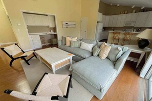 Ein Wohnzimmer mit einer Couch, einem Couchtisch und einem Esstisch. Es gibt eine Küchenzeile mit Schränken und einer Mikrowelle.
