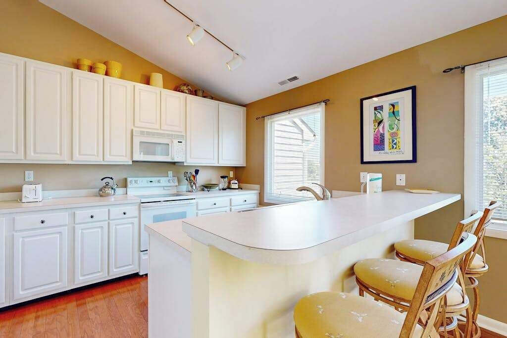 A kitchen with white cabinets, a central island with bar stools, a microwave, and a window with blinds.
