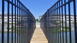Un chemin en bois, bordé de rampes métalliques, mène à un bâtiment doté d’une piscine.