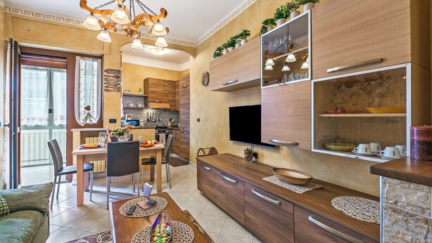 Een moderne keuken met houten kastjes, een eethoek met tafel en stoelen, een tv boven de aanrecht en een kroonluchter.