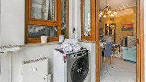 Een wasruimte met een wasmachine, een eettafel met stoelen en een bank in een kamer met houten ramen en een kroonluchter.