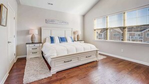 Une chambre avec un grand lit, deux tables de chevet, un tapis blanc, un ventilateur de plafond et une fenêtre avec des stores.