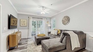 A living room with a grey sofa, ottoman, and a wooden side table. - The Tides spacious condo w/shared pool, close to restaurants & shopping (Rehoboth Beach)