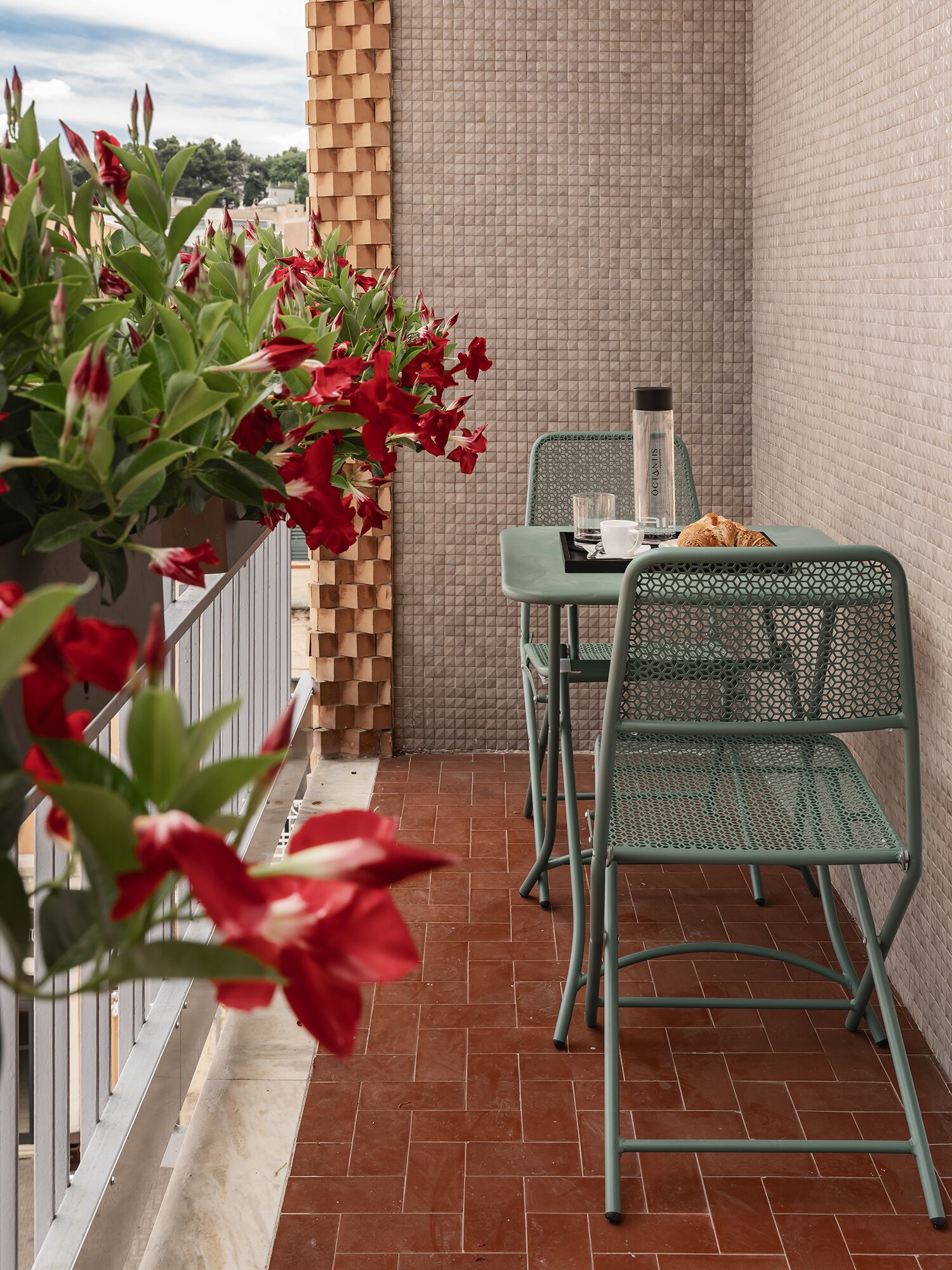 Varanda com mesa e cadeiras, um vaso com flores vermelhas e uma parede texturizada.
