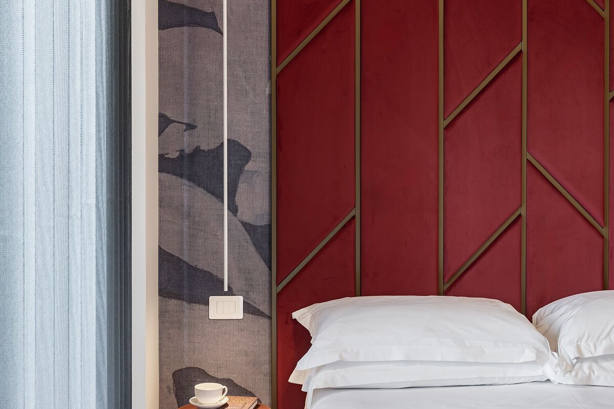 A hotel room with a bed, a side table with a book and a cup, a red accent wall, and a patterned tapestry.