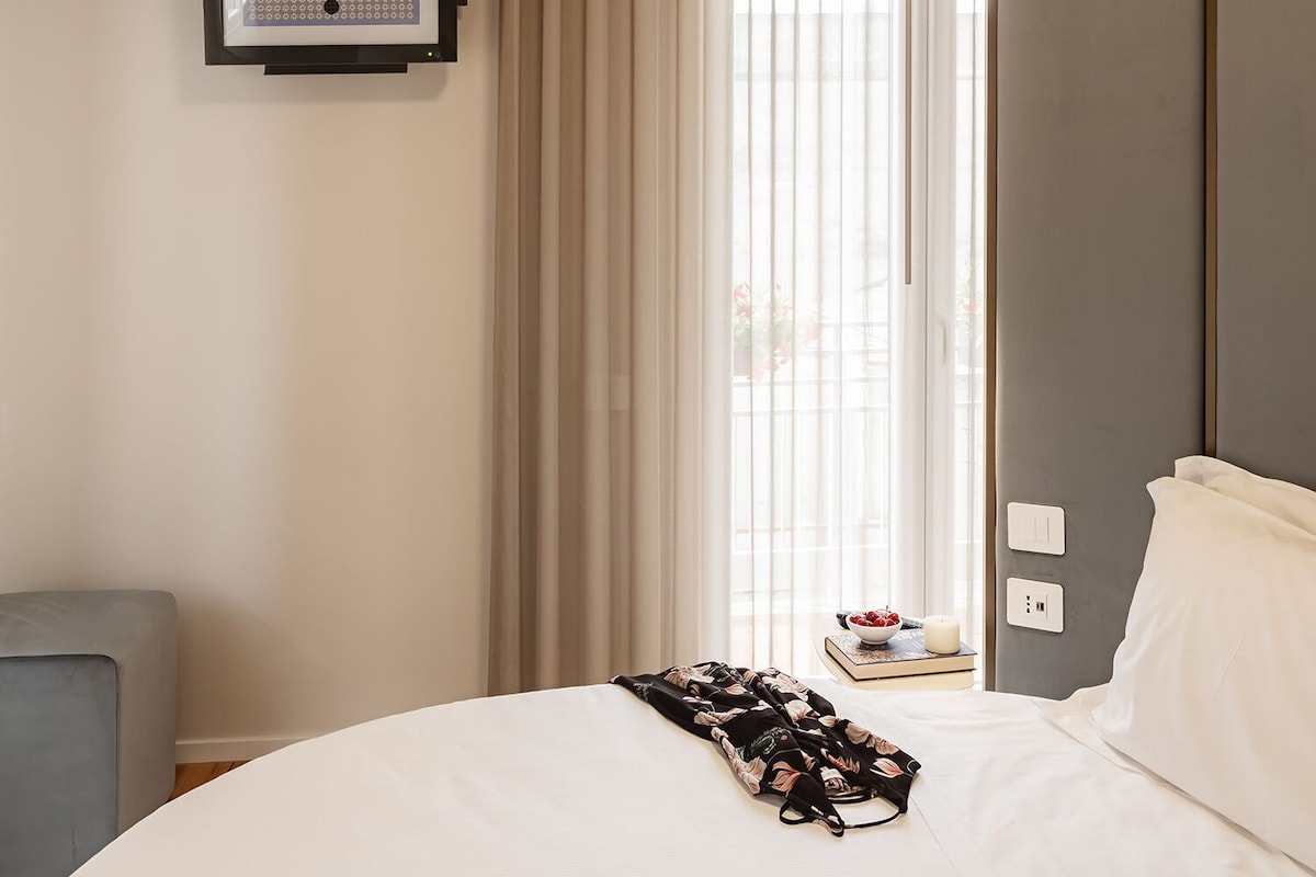 A hotel room with a bed, a bedside table with a bowl of fruit, a framed picture on the wall, and a window with curtains.