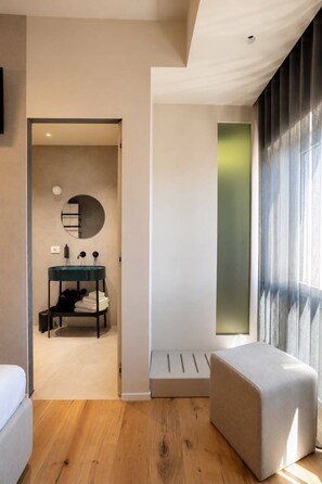 A modern room with a wooden floor, a round mirror, a small table with a glass top, a bench, and a large window with curtains.